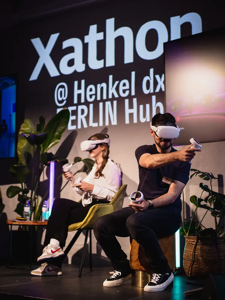 A man and a women are sitting on stage holding a remote control and wearing VR glasses.